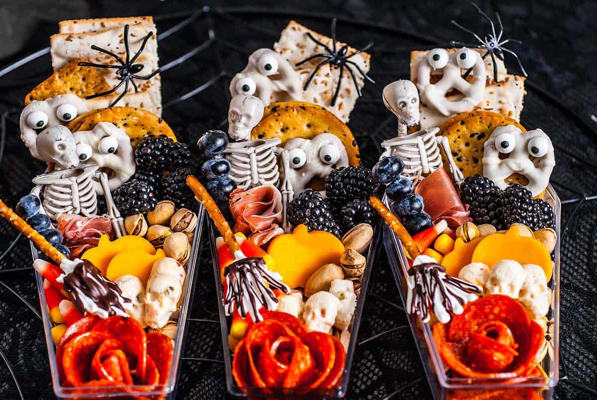 Three snack cups with crackers, cheese, meats, berries, nuts, skeleton and ghost picks, and plastic spider toppers.