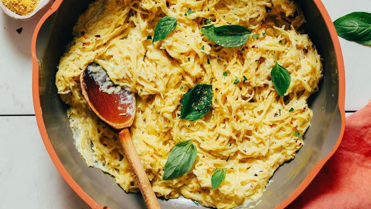 A skillet filled with creamy spaghetti squash topped with fresh basil leaves, with a wooden spoon resting inside.