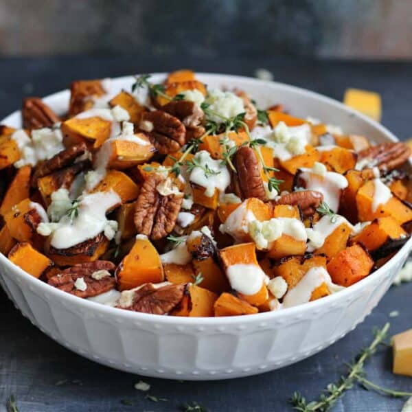 A white bowl with roasted butternut squash cubes, toasted pecans, melted cheese, and fresh herbs on a dark surface.