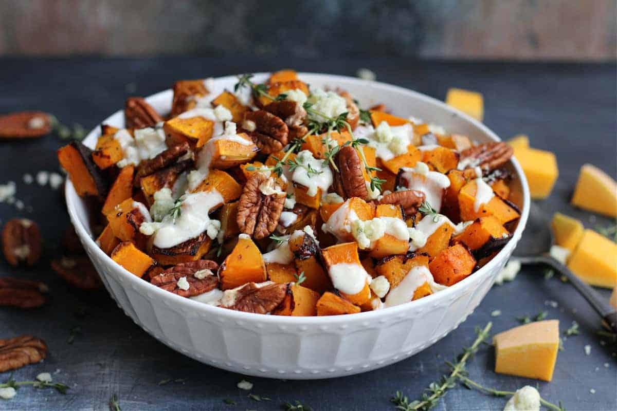 A white bowl with roasted butternut squash cubes, toasted pecans, melted cheese, and fresh herbs on a dark surface.