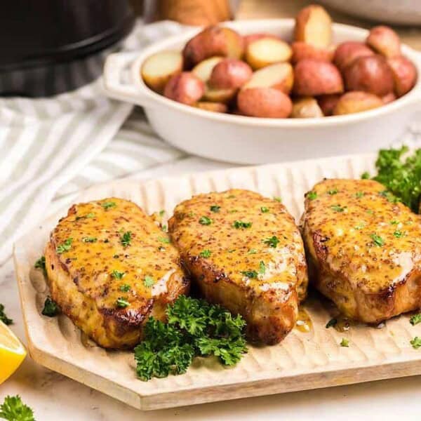 Four glazed pork chops with parsley on a wooden platter, a bowl of roasted potatoes, and a halved lemon in the background.