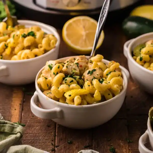 Three white bowls of creamy pasta with herbs on a wooden table; zucchini, lemons, and a slow cooker are in the background.