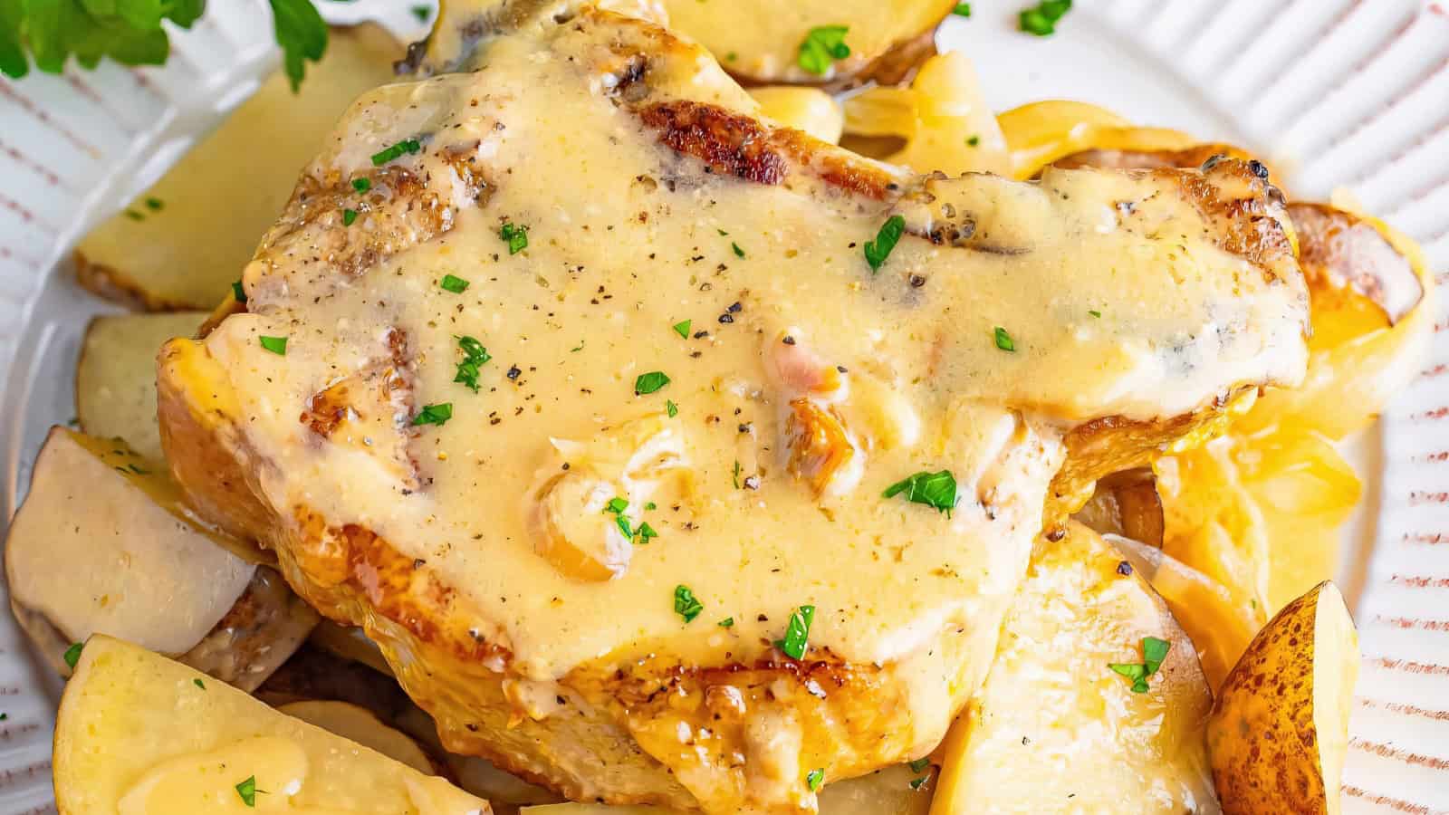 A plate featuring a grilled pork chop topped with creamy garlic sauce and garnished with chopped parsley. Sliced potatoes are arranged around the main dish. The meal is presented on a white plate with a decorative edge.