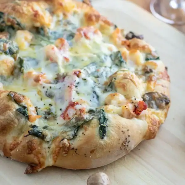 A baked pizza with melted cheese, spinach, and seafood sits on a wooden board with whole mushrooms placed beside it.