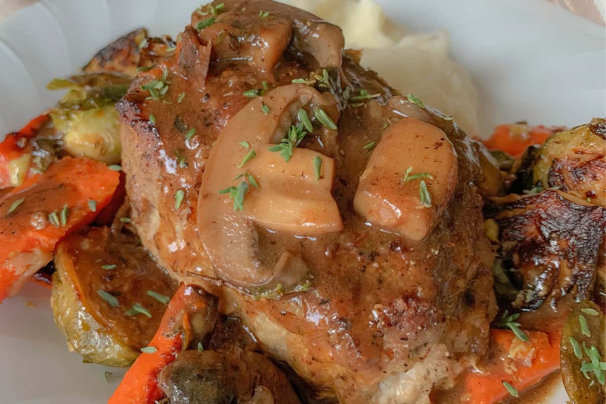 A plate featuring a succulent beef steak topped with a rich mushroom sauce, garnished with fresh herbs. Surrounding the steak are roasted vegetables, including carrots and brussels sprouts, resting on a bed of creamy mashed potatoes.