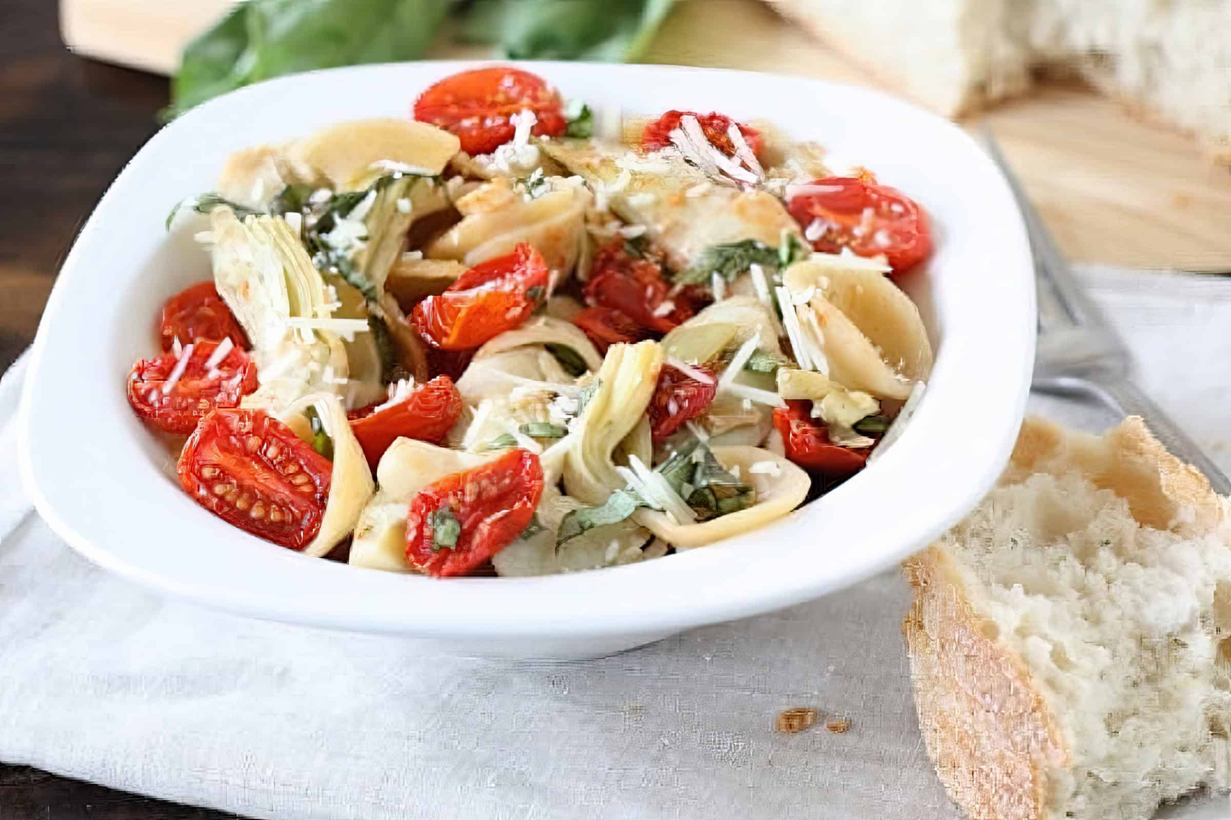 A white bowl filled with pasta, cherry tomatoes, artichoke hearts, and grated cheese, garnished with fresh basil. A piece of crusty bread is beside the bowl on a light-colored cloth.