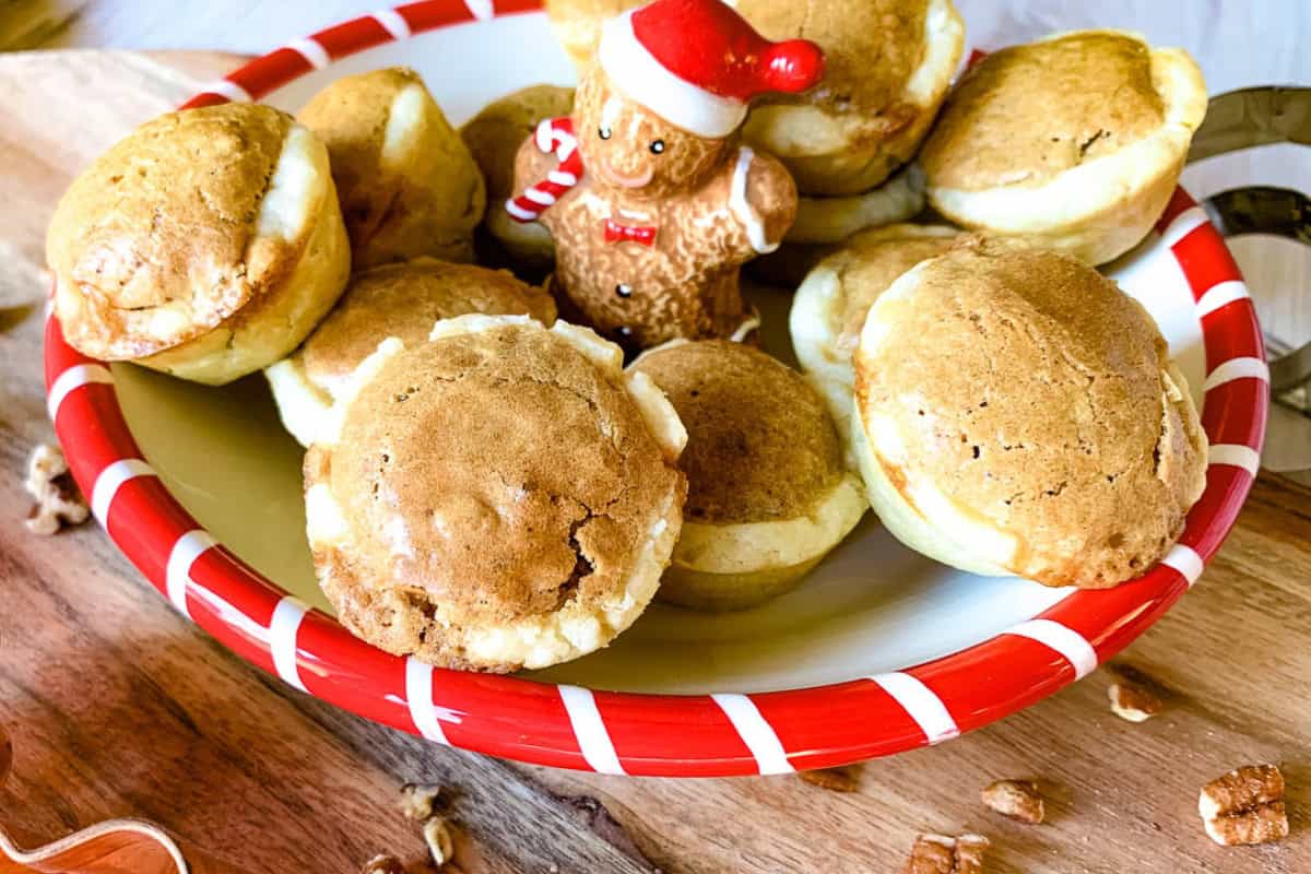 A festive plate with cookies topped with a small gingerbread man wearing a Santa hat. The plate has a red and white stripe pattern, and it sits on a wooden surface sprinkled with nuts.