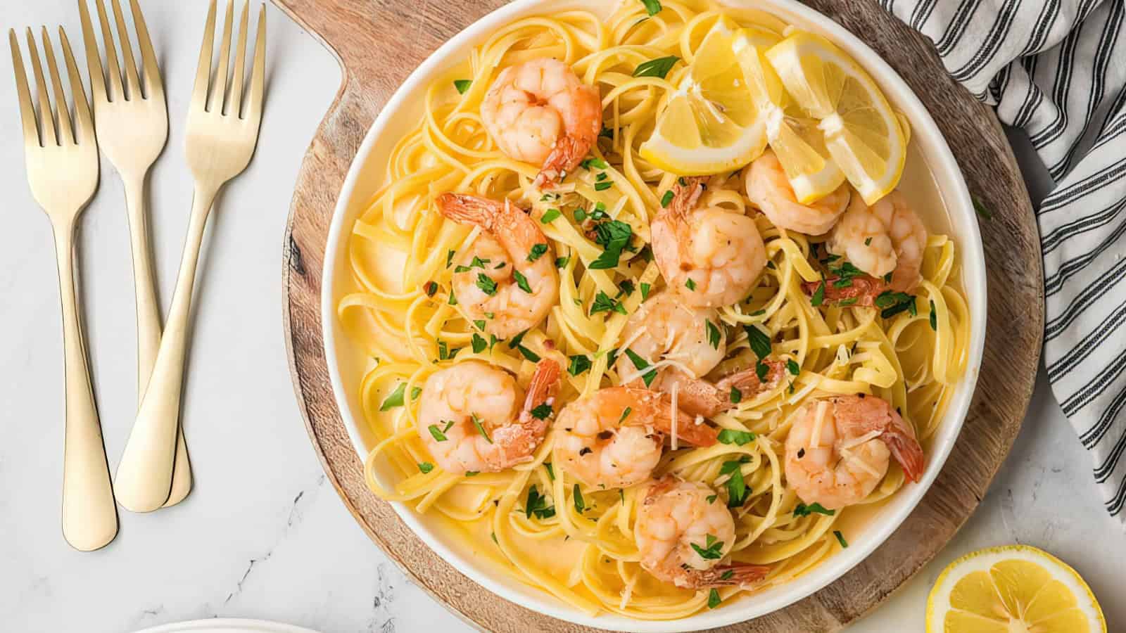 A plate of shrimp scampi pasta garnished with parsley and lemon slices sits on a wooden board. Three gold forks are beside it, with a striped napkin nearby. The background features a marble surface.