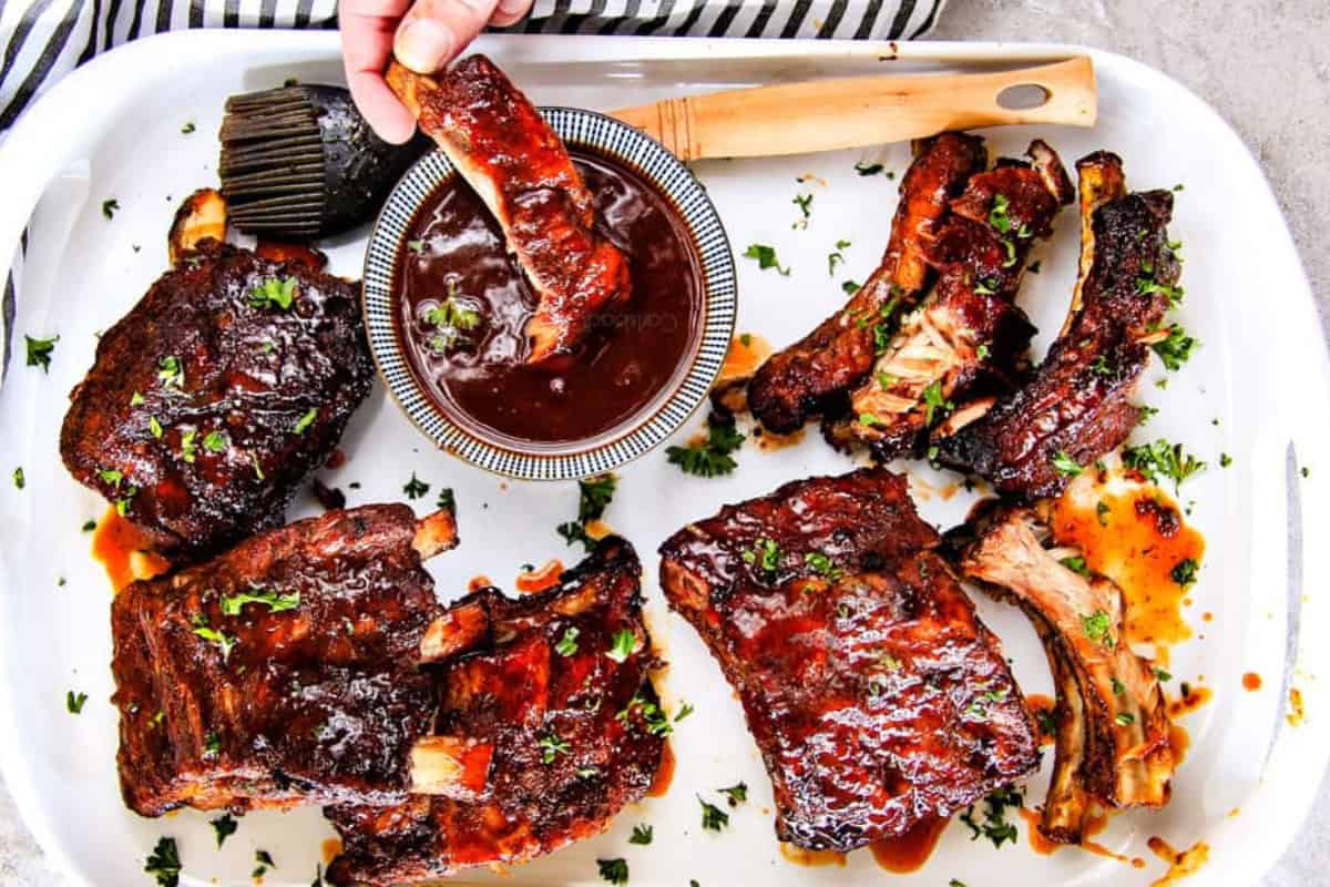 A platter of glazed barbecue ribs garnished with herbs. A hand dips a rib into a small bowl of barbecue sauce. A pastry brush with sauce is placed nearby.