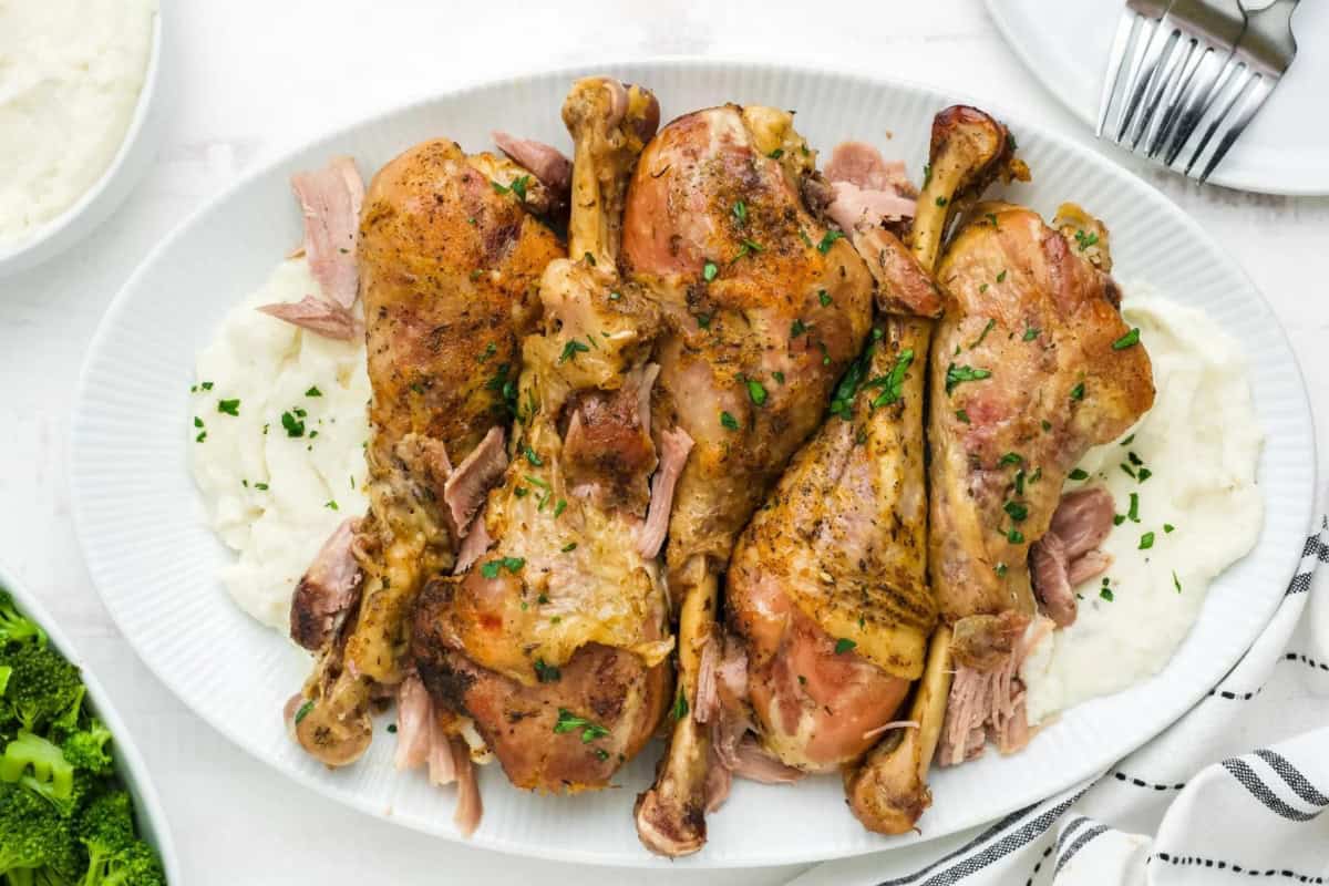 A platter of cooked turkey legs garnished with herbs, placed on a bed of creamy mashed potatoes. A side dish of broccoli and a partial view of a cutlery set are visible in the background.