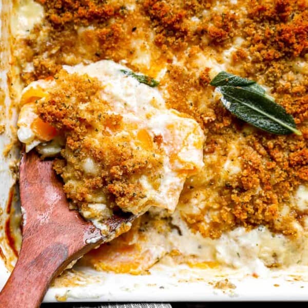A close-up of a breadcrumb-topped gratin in a casserole dish with a serving on a wooden spoon, garnished with sage leaves.