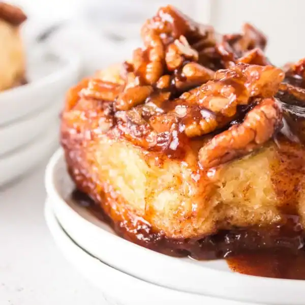 A close-up of a sticky bun with chopped pecans and caramel sauce on a white plate.