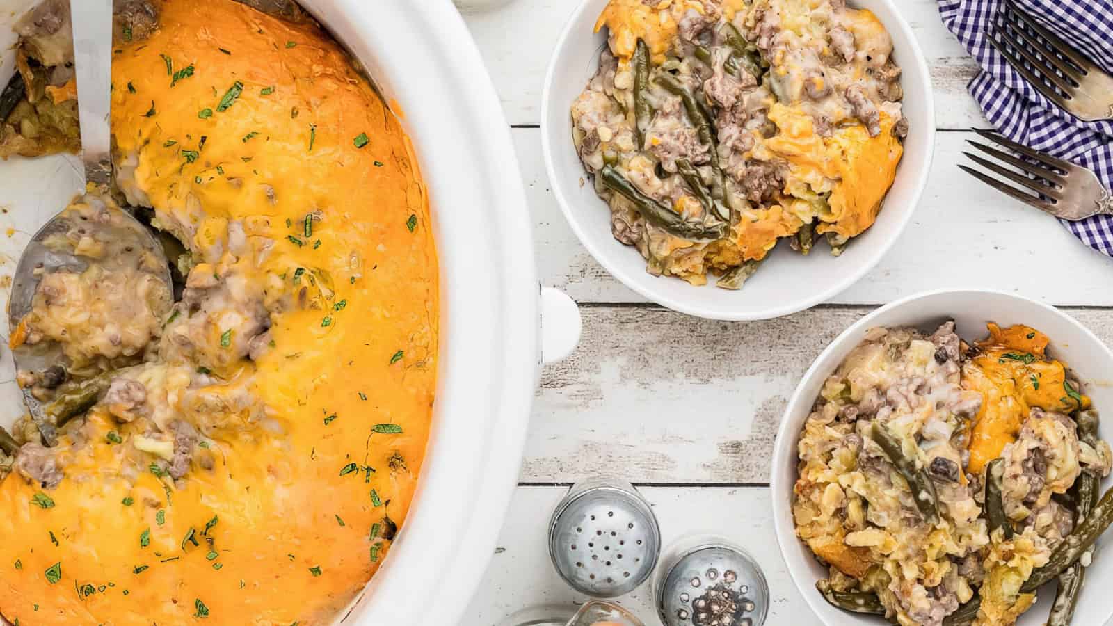 A close-up of two bowls filled with a cheesy beef and green bean casserole, along with a serving in a slow cooker. The casserole is topped with melted cheese. Fork, spoon, and a blue checkered napkin are visible on a rustic white table.