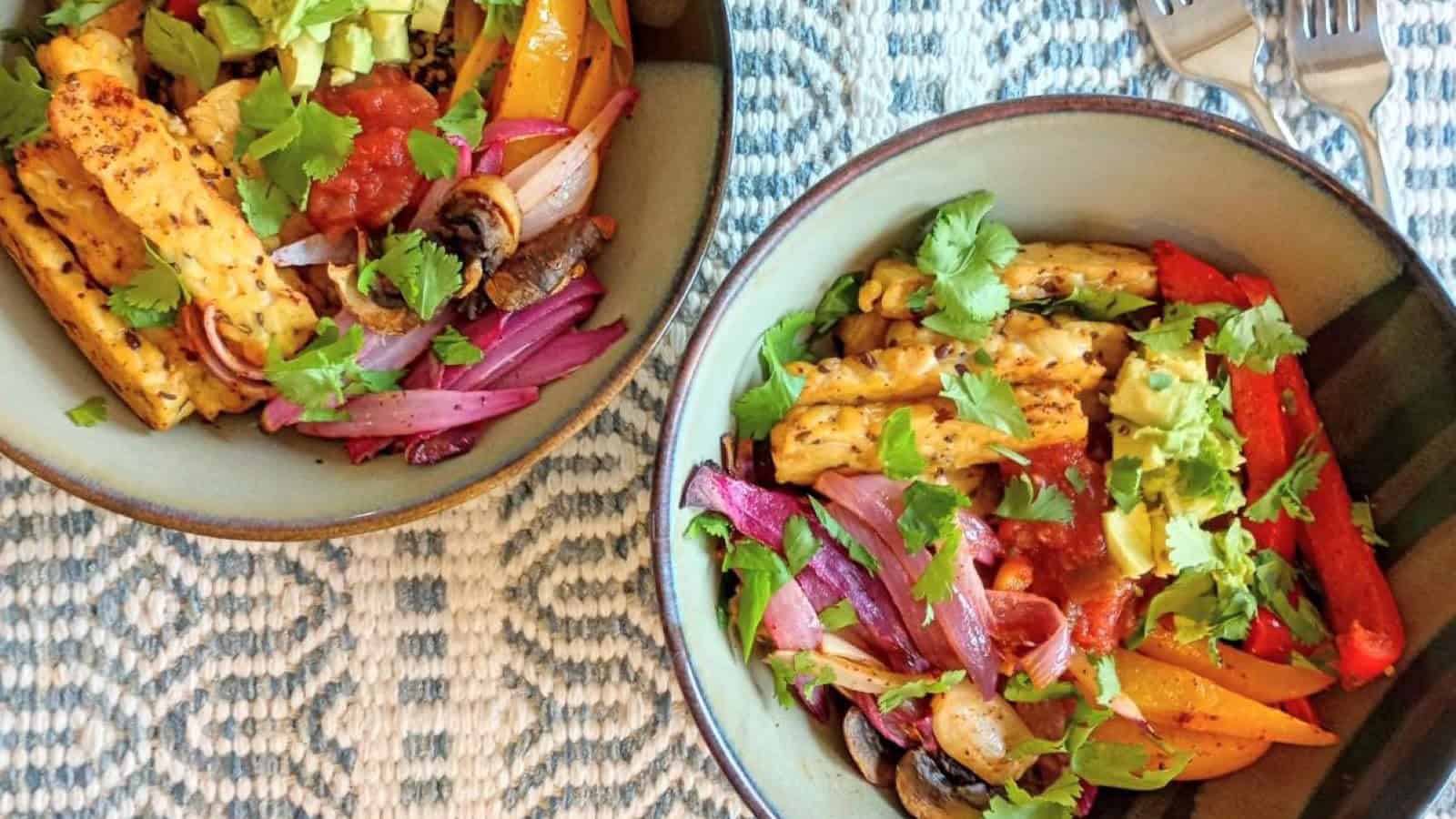 Two bowls of vibrant vegetable fajitas with red onion, bell peppers, zucchini, avocado, mushrooms, and cilantro on a blue patterned tablecloth, accompanied by a fork on the side.