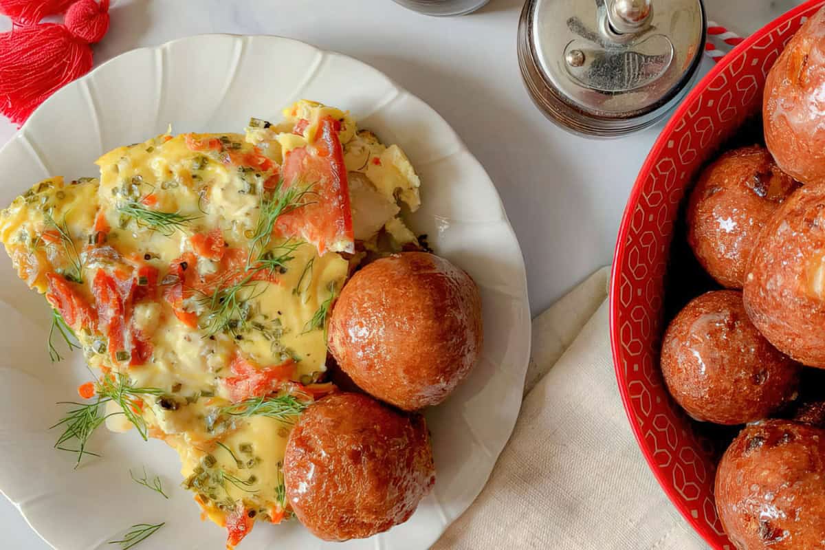 A plate with a slice of omelette topped with herbs and sliced red peppers, accompanied by two round, golden-brown fried balls. A red bowl filled with more fried balls and pepper shakers are nearby on a white surface.