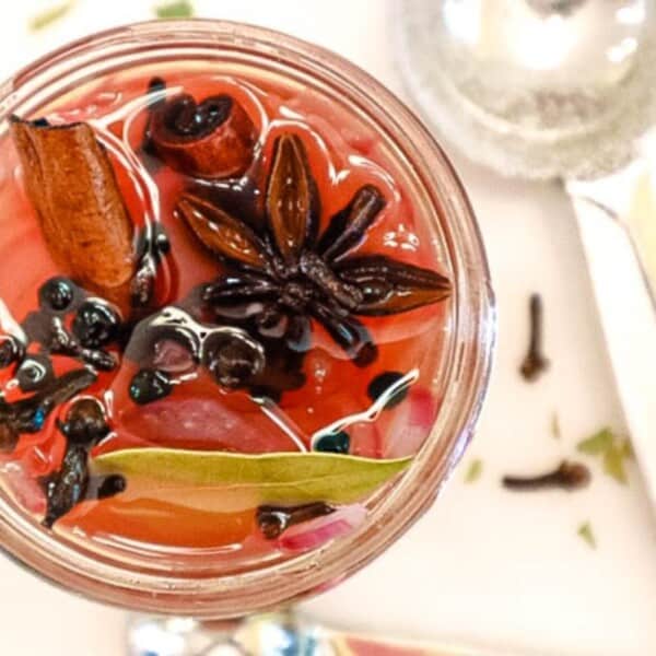 A glass jar of pickling brine containing star anise, cinnamon sticks, bay leaves, and whole peppercorns.