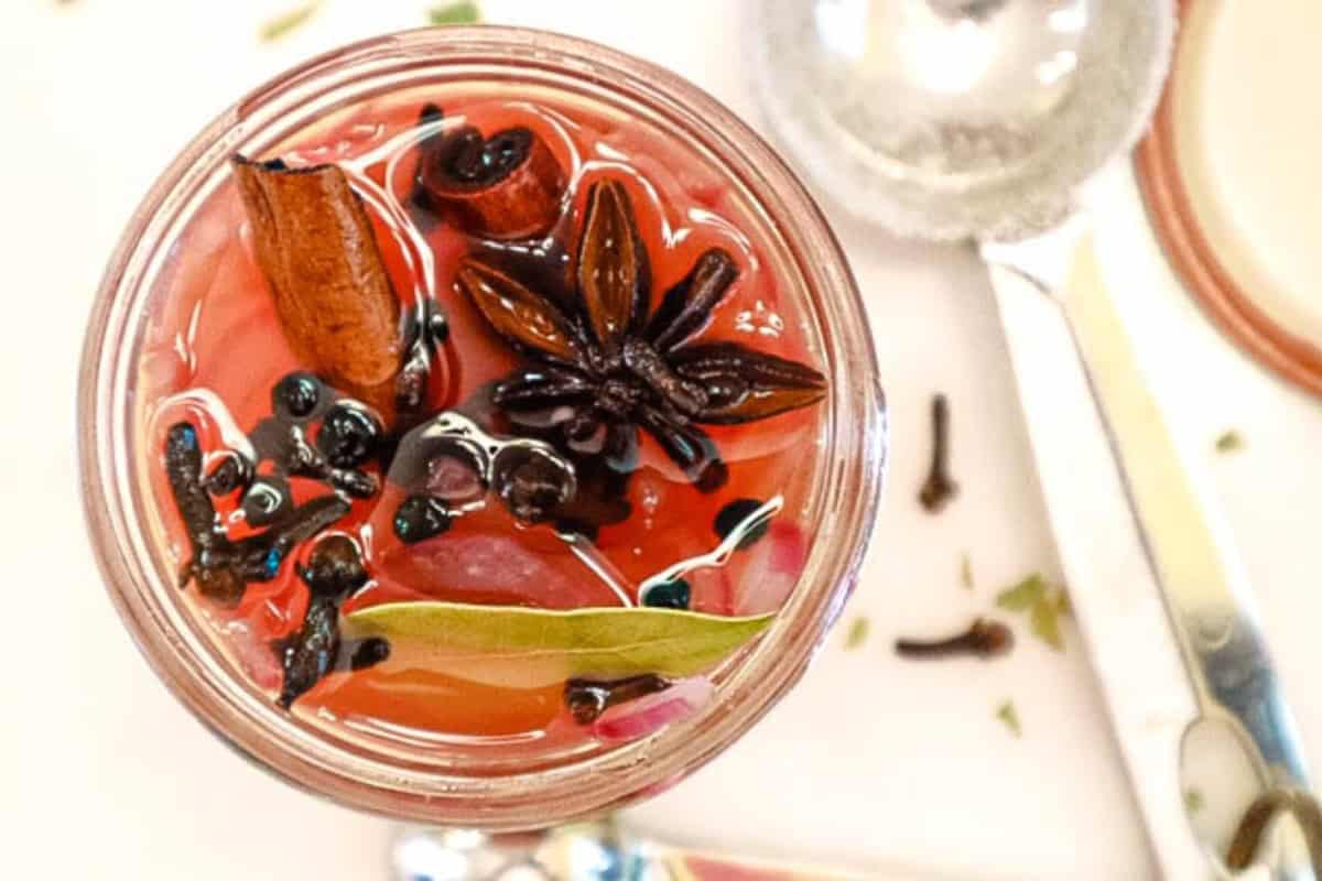 A close-up of a jar filled with a red liquid, featuring spices like cinnamon sticks, star anise, bay leaves, and cloves. A spoon lies next to the jar on a white surface.
