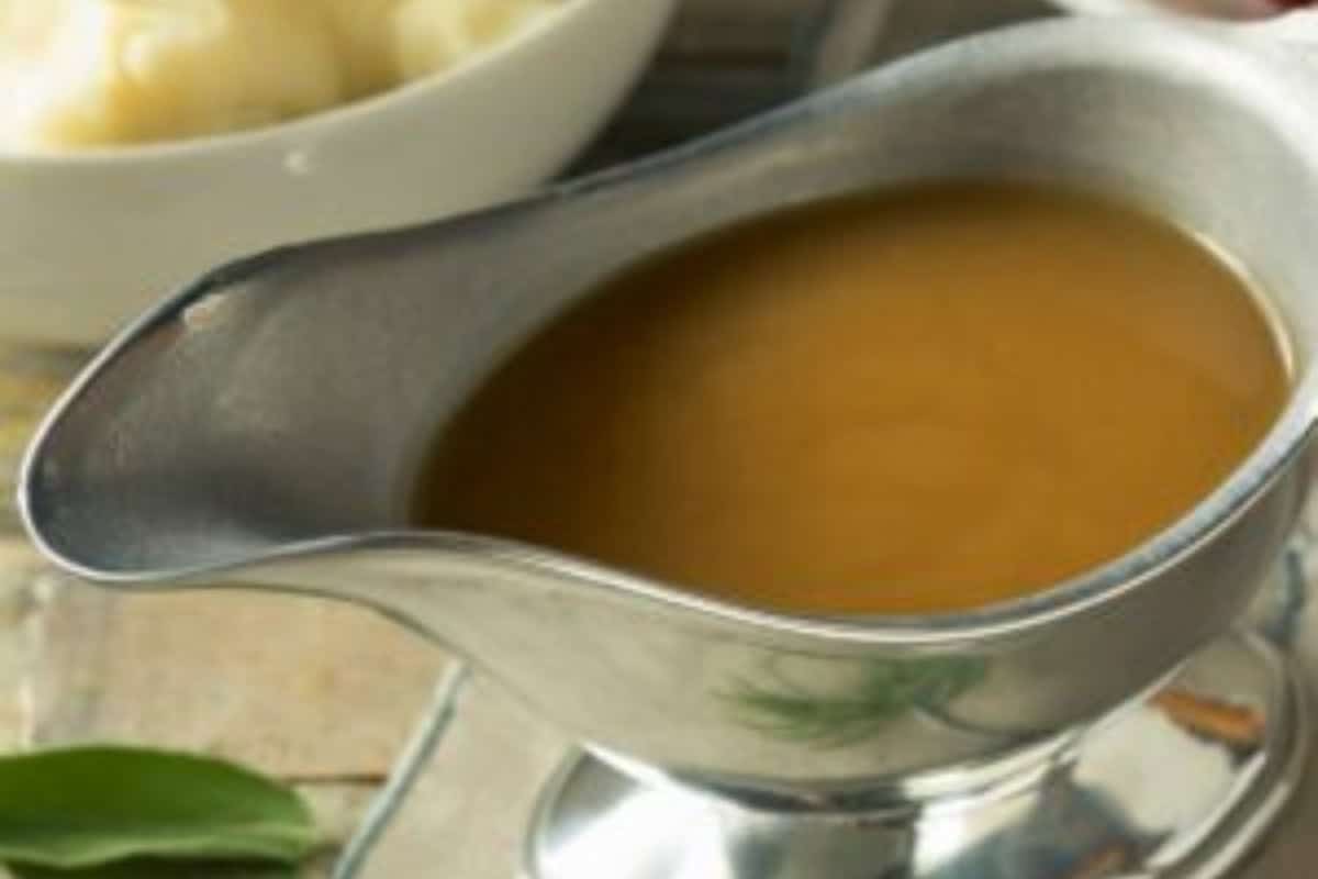 A silver gravy boat filled with brown gravy sits on a table. In the background, there is a bowl of mashed potatoes.