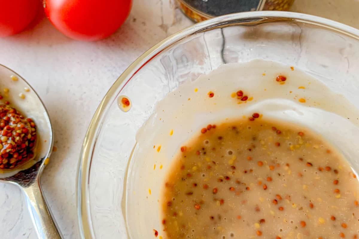 A glass bowl containing a creamy mustard dressing with mustard seeds, beside a spoon holding mustard. Two ripe tomatoes are partially visible in the background on a light surface.