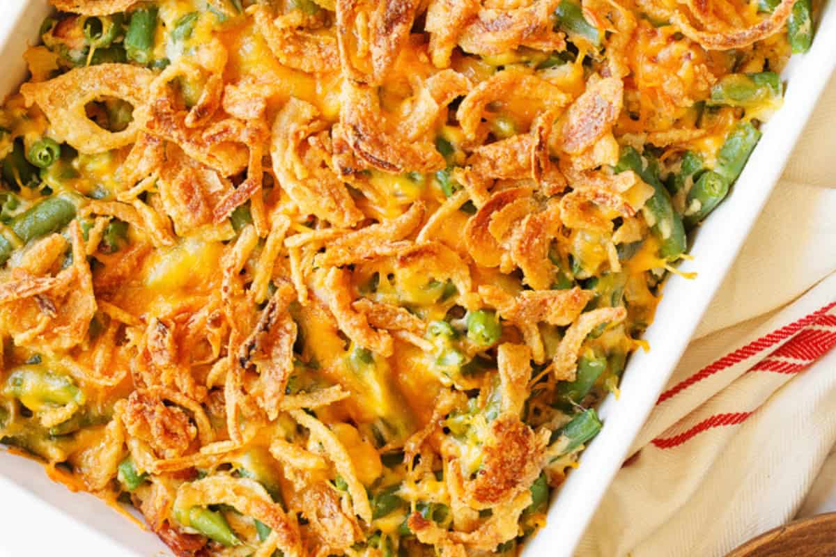 A baked green bean casserole topped with crispy fried onions and melted cheese in a white rectangular dish. The dish is placed on a striped red and white cloth.