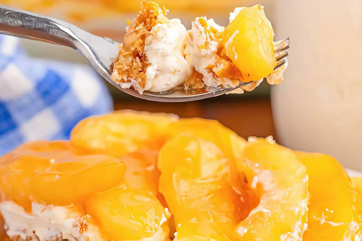 A fork holds a bite of dessert with peach slices, creamy filling, and crumbly topping. Below, more peach slices and creamy filling are visible. A blue and white checkered cloth is in the background.