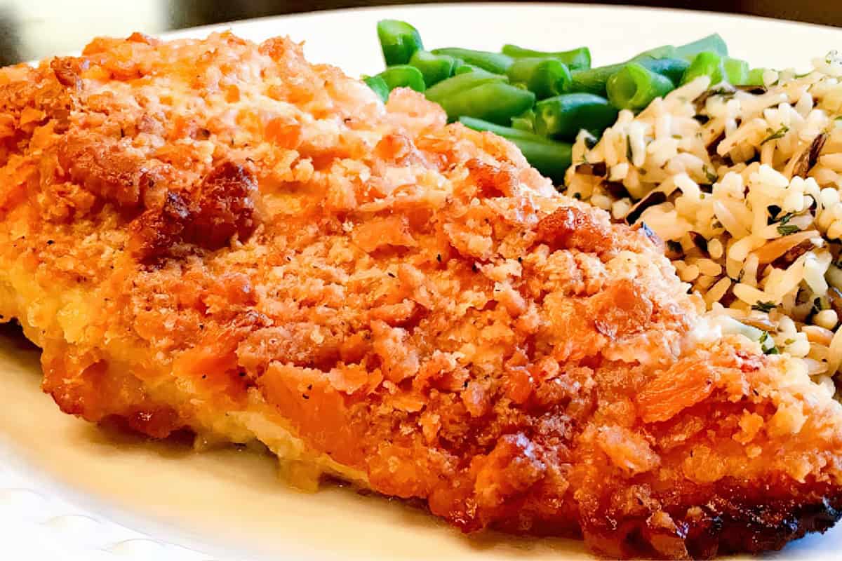 A crispy baked chicken breast is served on a white plate, accompanied by green beans and a portion of rice mixed with herbs.