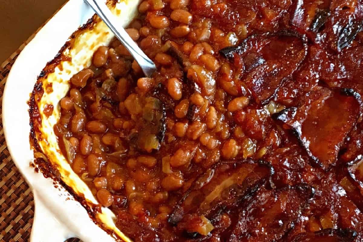 A baked dish of beans topped with slices of browned, crispy bacon, served in a white casserole dish with a spoon. The surface is slightly caramelized around the edges.
