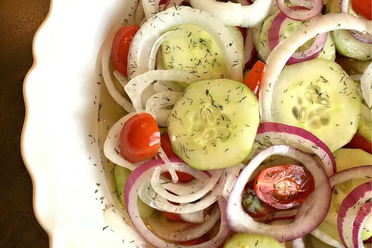 A close-up of a salad in a white bowl, featuring slices of cucumber, red onion, and cherry tomatoes, sprinkled with dill. The fresh ingredients create a colorful, appetizing display.