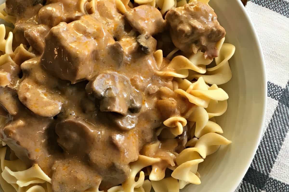 A plate of creamy beef stroganoff with wide egg noodles. The rich sauce covers chunks of beef and mushrooms, served on a light-colored dish. A checkered napkin is partially visible next to the plate.