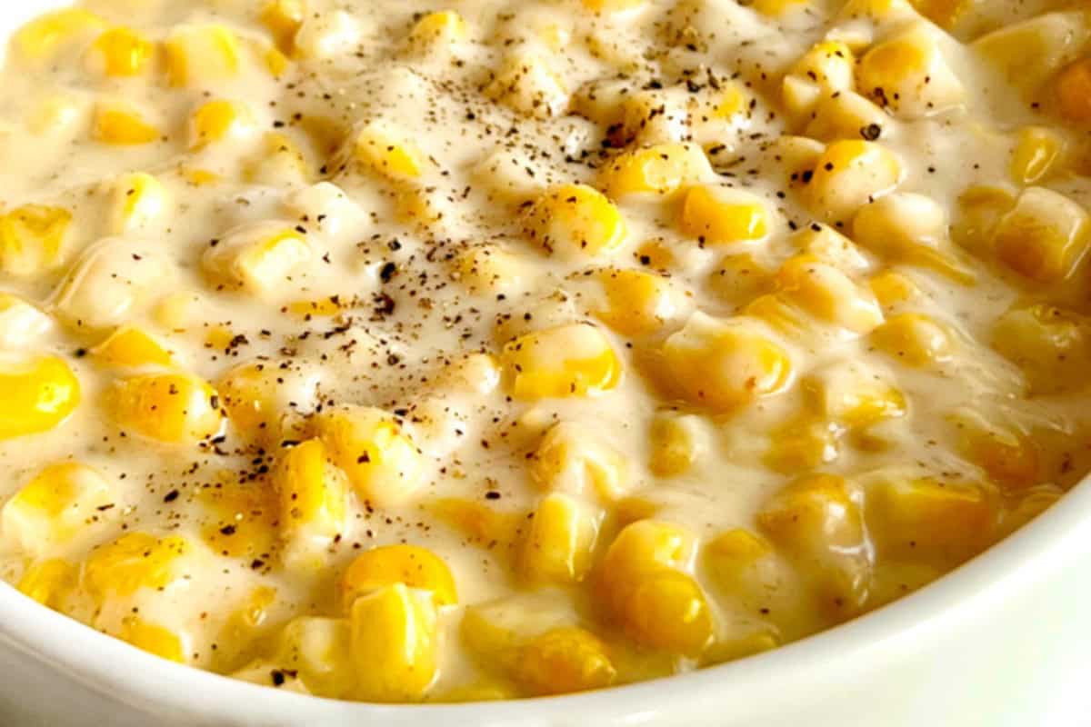 A close-up of creamy corn in a white bowl, topped with a sprinkle of black pepper. The corn appears to be in a thick, creamy sauce.