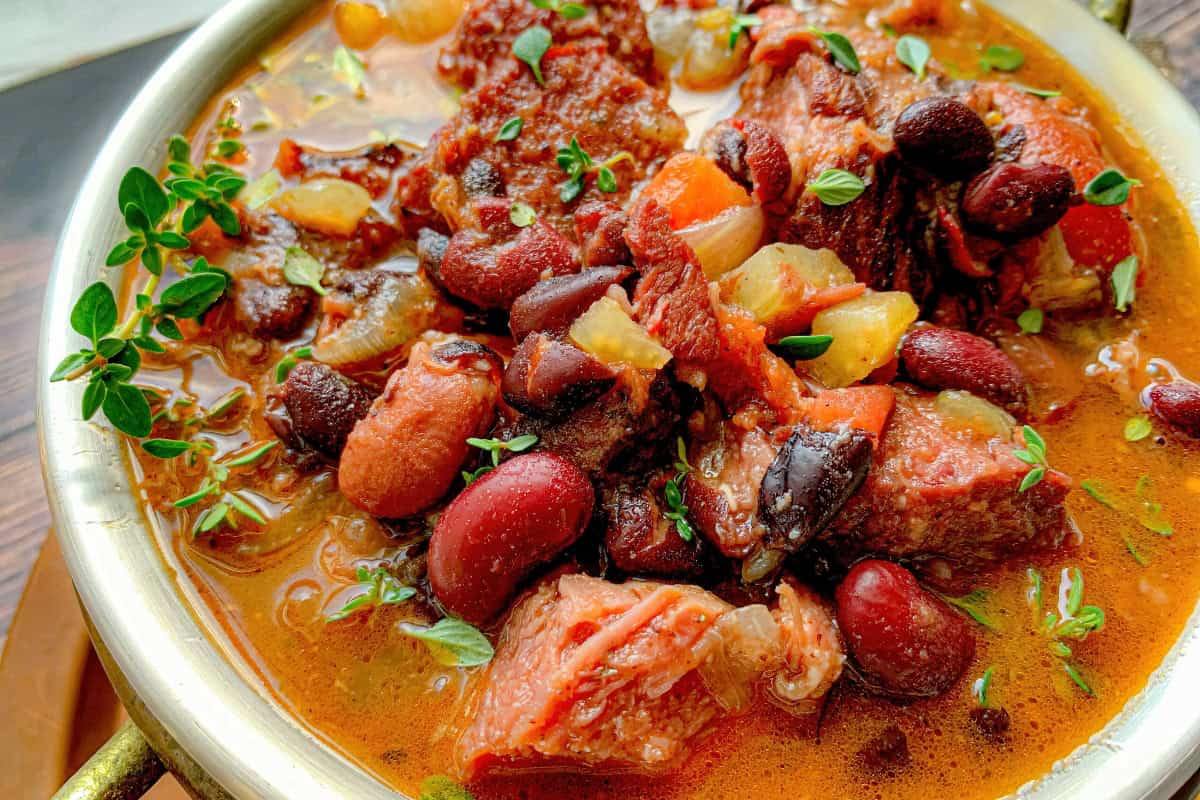 A close-up of a hearty beef stew in a bowl, featuring chunks of beef, mixed beans, and vegetables. It is garnished with fresh herbs and has a rich, colorful broth.