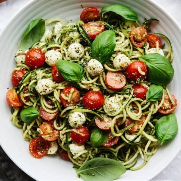 A white plate with zucchini noodles, cherry tomatoes, mozzarella balls, fresh basil leaves, and a sprinkle of pepper.