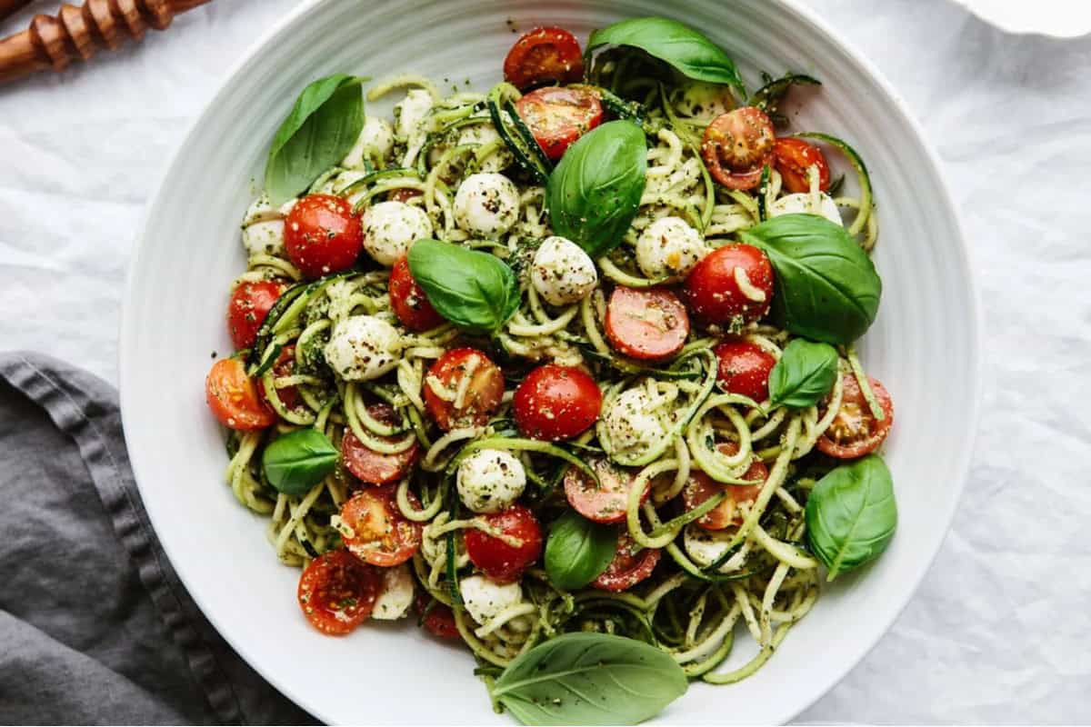 A white plate filled with zucchini noodles, cherry tomatoes, mozzarella balls, and fresh basil leaves, all seasoned with black pepper. The dish is placed on a white surface with a gray cloth to the side.