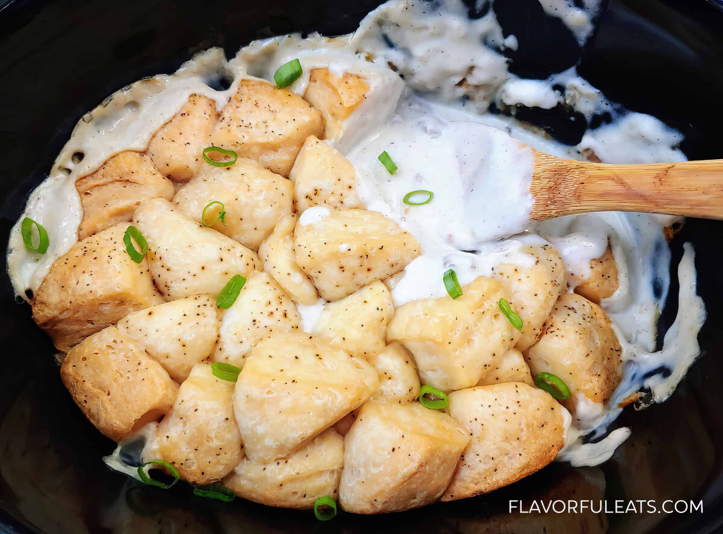 The image shows golden biscuits topped with creamy sauce and chopped green onions in a slow cooker, accompanied by a wooden spoon.