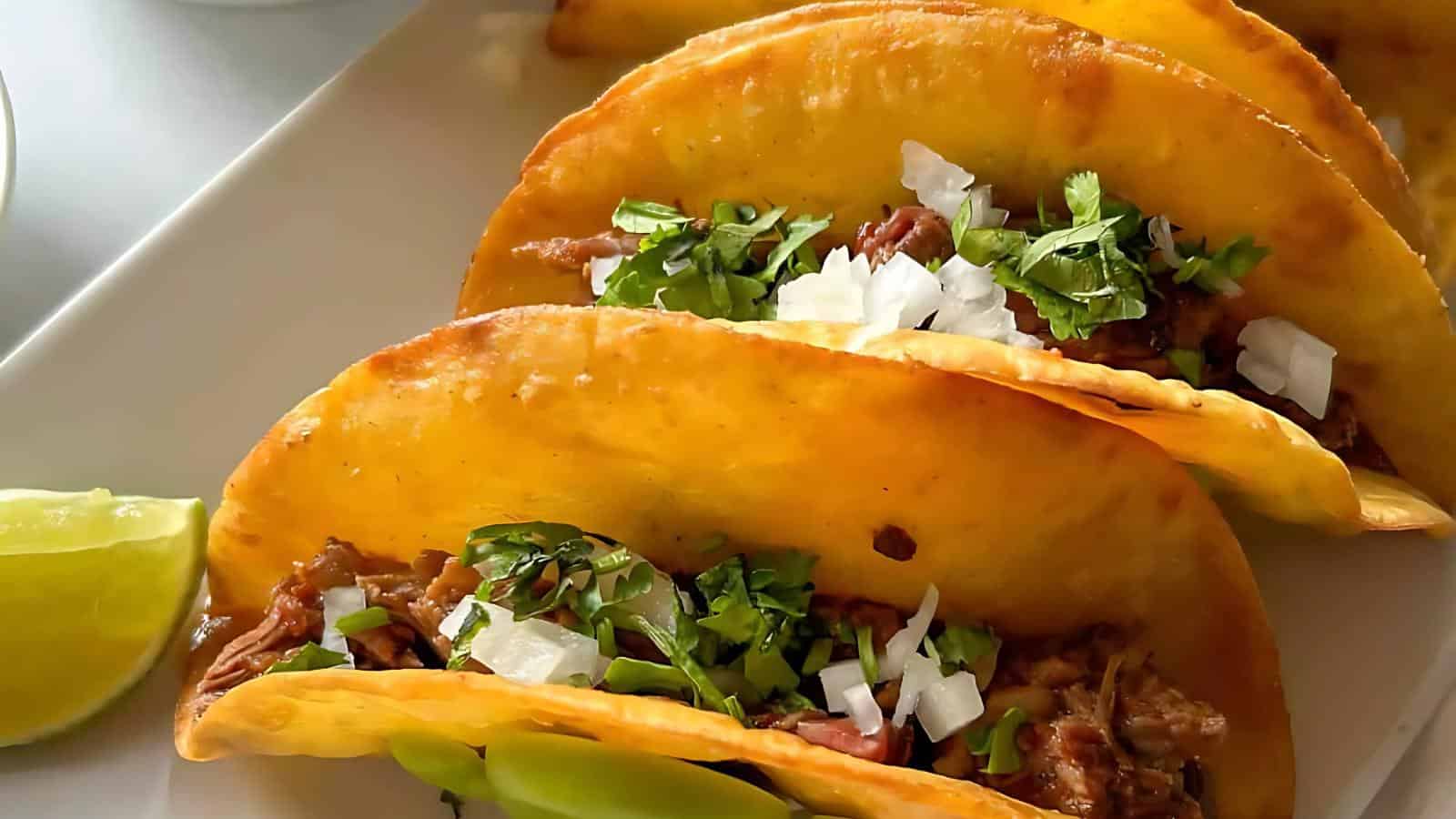 A plate holds three tacos filled with meat, cilantro, and onions, accompanied by lime wedges.