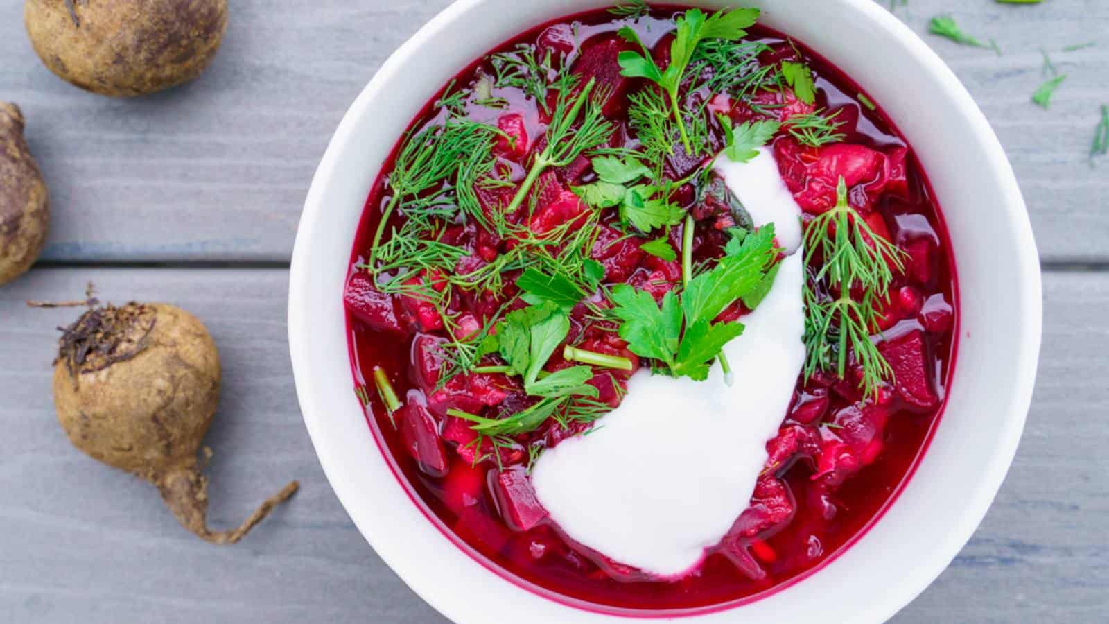 A bowl of red beet soup with a dollop of sour cream and fresh herbs, placed next to whole beets on a gray wooden table.
