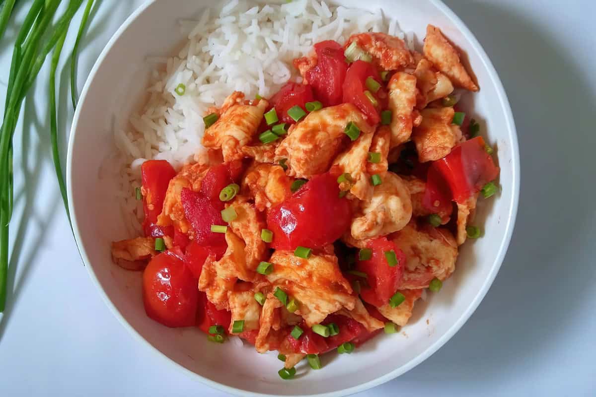 A bowl of white rice with stir-fried chicken, tomatoes, and chopped green onions on top. Fresh green onions are placed beside the bowl.