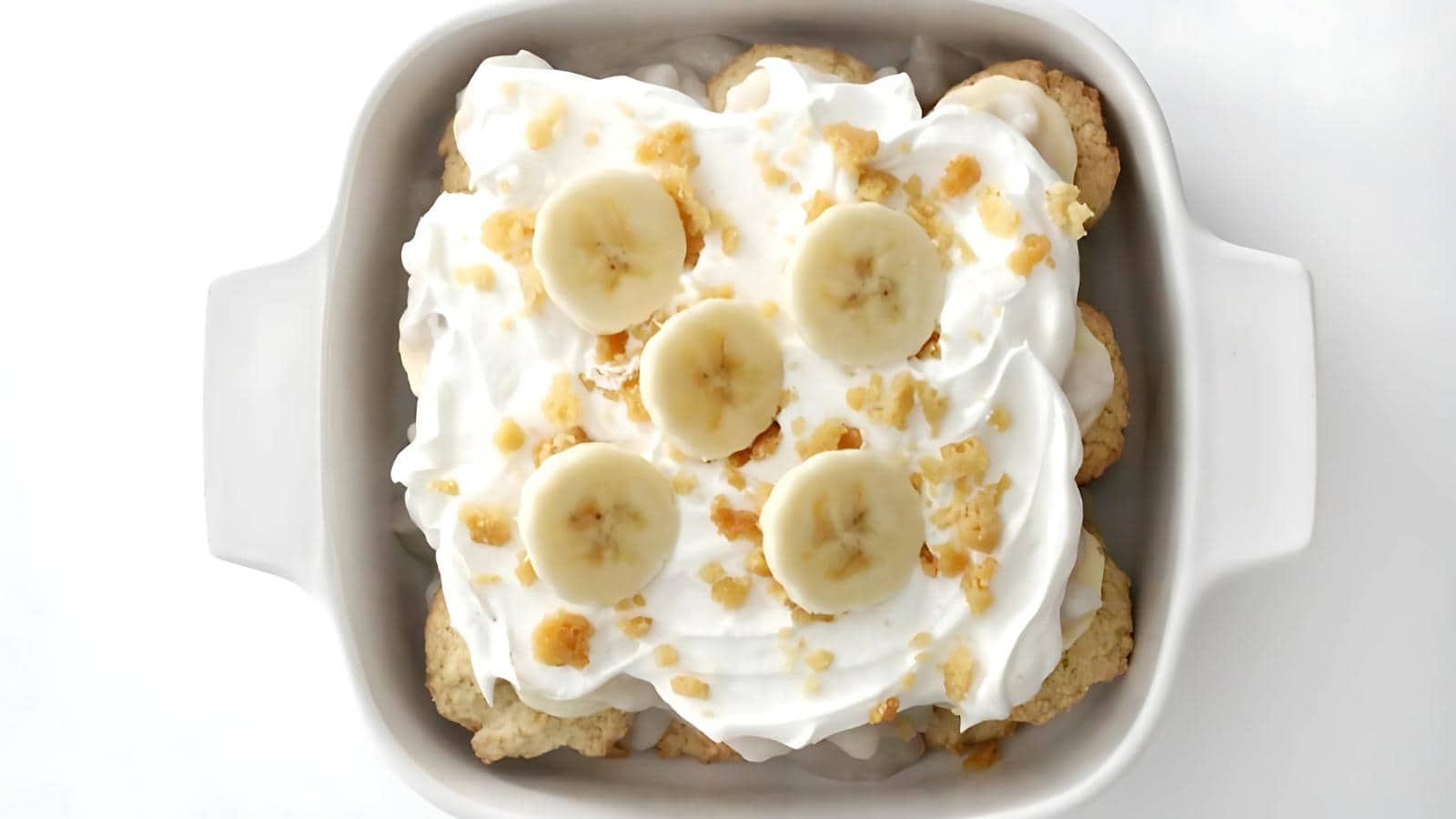 A dessert dish topped with sliced bananas, whipped cream, and crumbled nuts, served in a square white baking dish.
