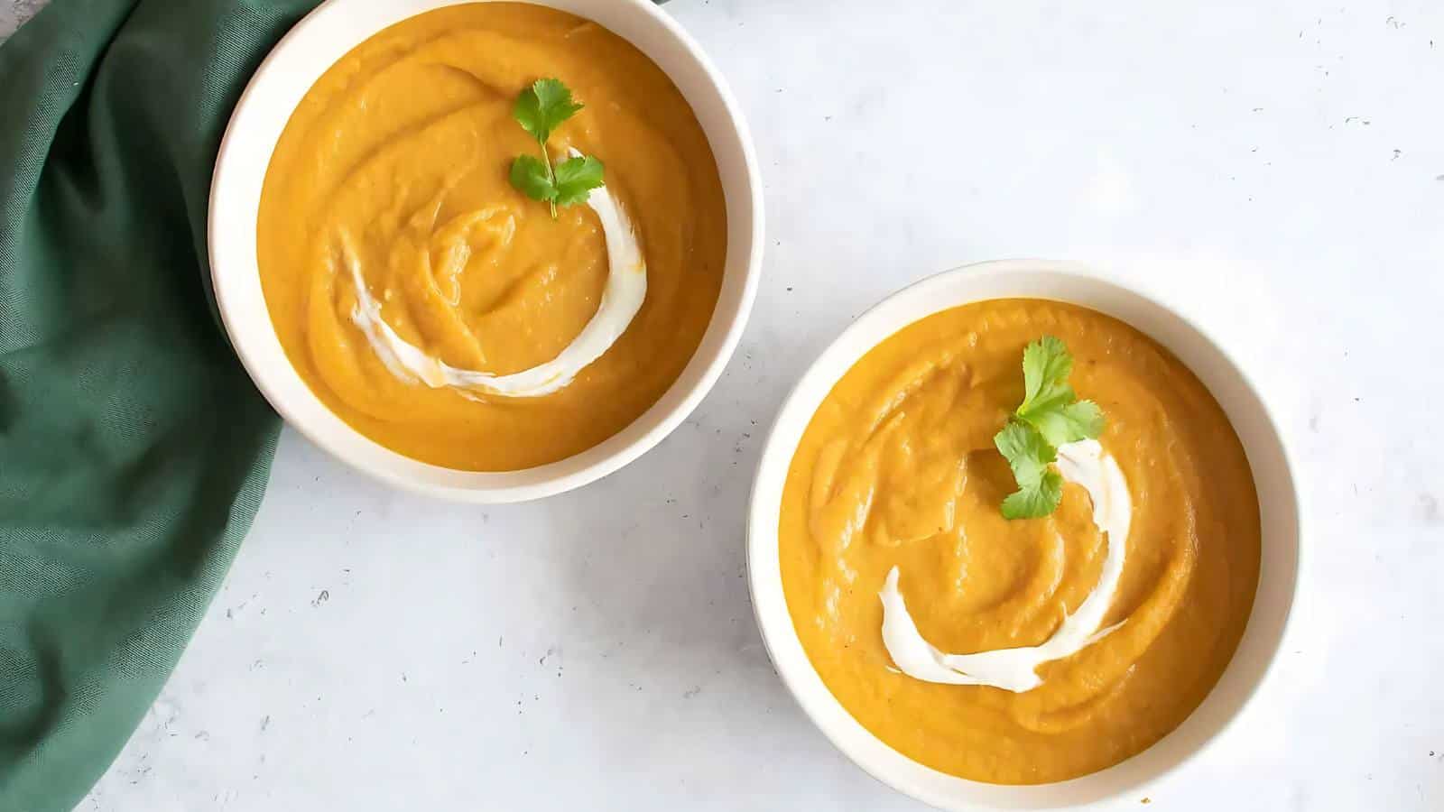 Two bowls of orange soup topped with cream and cilantro on a white surface, with a green cloth nearby.