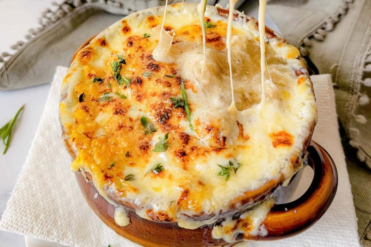 A baked dish of French onion soup topped with melted and browned cheese, garnished with fresh herbs. The cheese is stringy, pulling away from the soup, served in a brown ceramic bowl on a white napkin.