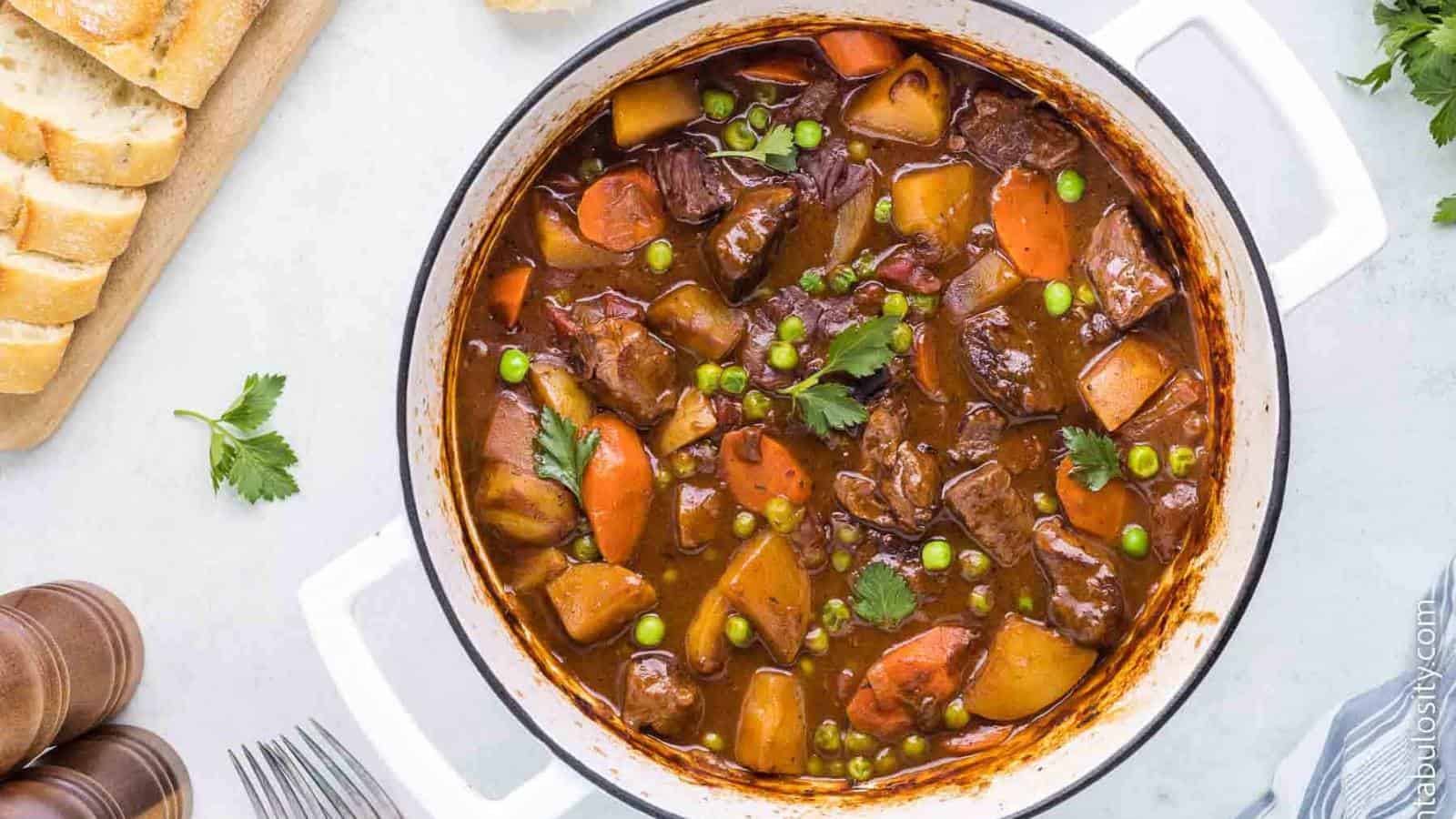 A pot containing chunks of meat, potatoes, carrots, and peas in a rich broth. Nearby, slices of bread are placed on a cutting board.