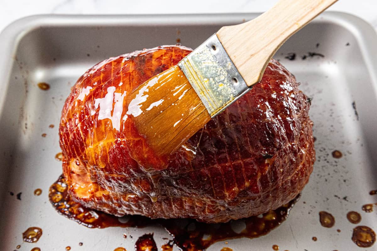A brown sugar glazed ham is being brushed with glaze in a baking pan. The ham has a shiny, caramelized finish, and the wooden brush is coated in glossy glaze, adding a rich layer to the meat.
