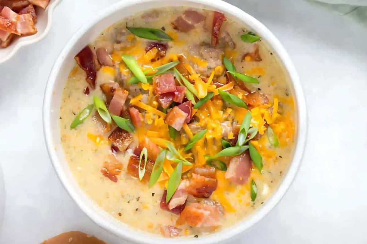 A bowl of creamy soup topped with chopped bacon, shredded cheddar cheese, and sliced green onions, presented on a white surface.