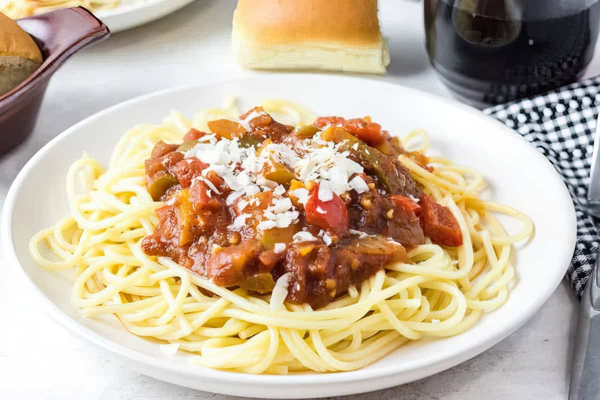 A plate of spaghetti topped with a chunky tomato sauce and grated cheese. The dish is accompanied by a bread roll in the background. A glass of red wine and checkered cloth are partially visible on the side.