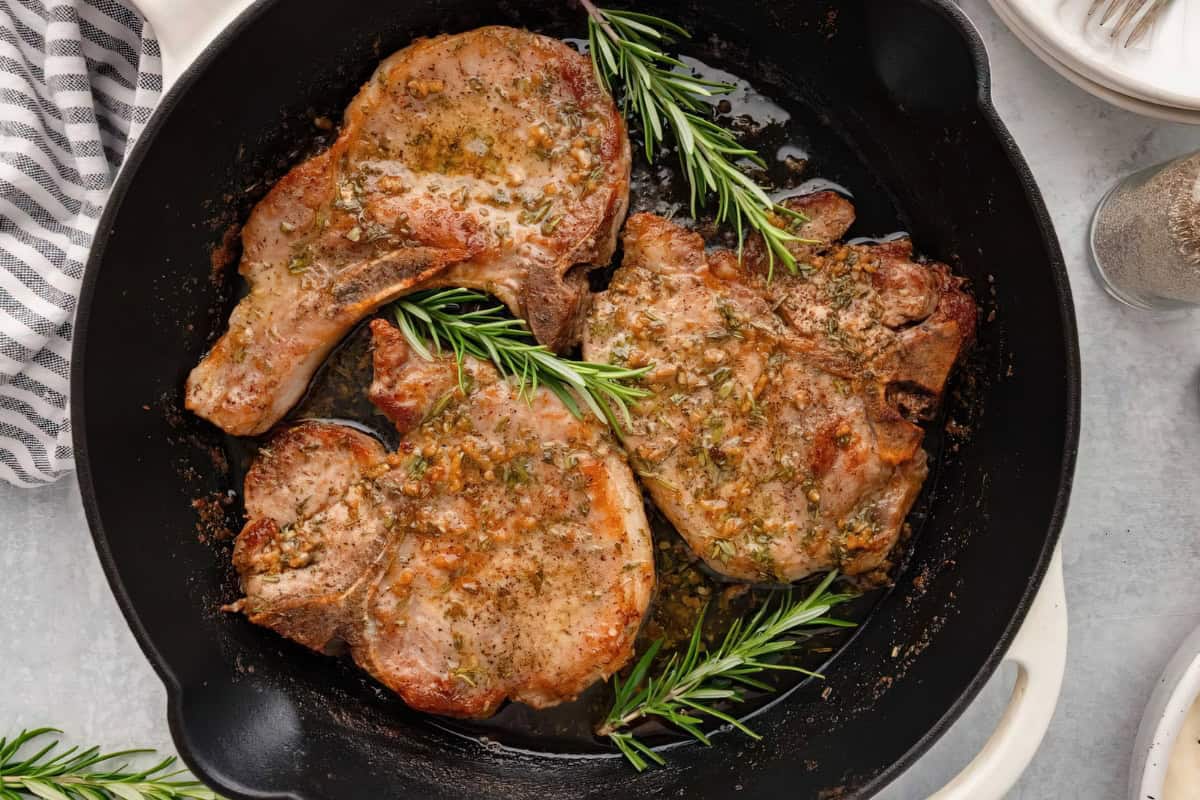 The image shows a black skillet containing three pork chops garnished with sprigs of rosemary.