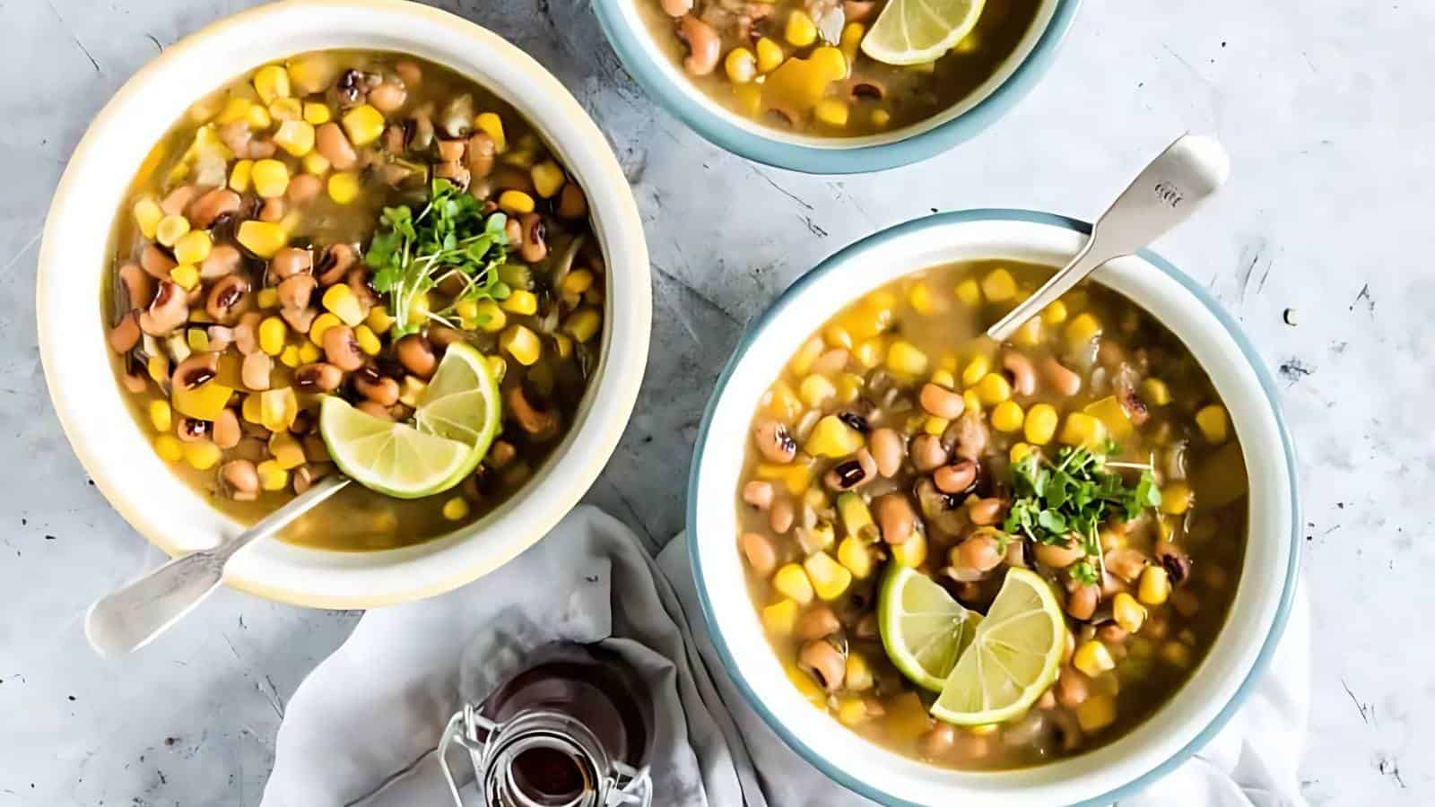 The image shows three bowls filled with bean and corn soup, each garnished with lime slices and herbs. A small glass jar is placed nearby on a light surface.