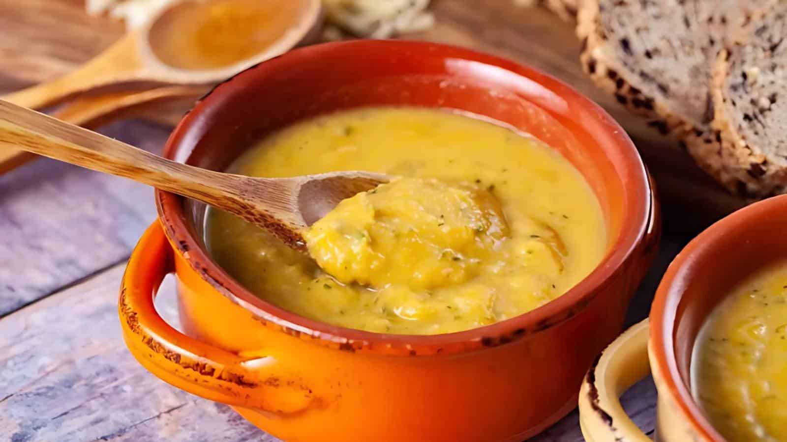 A wooden spoon in a bowl of thick, yellow soup placed in an orange ceramic dish. Slices of bread are visible in the background.