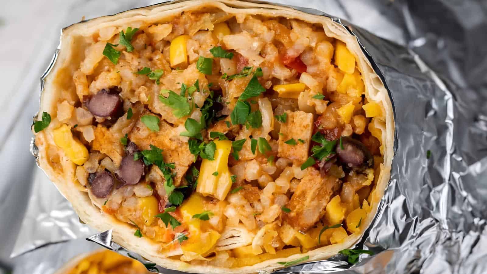 The image shows a burrito cut open, revealing a filling that includes rice, beans, corn, and cilantro. It is wrapped in foil.
