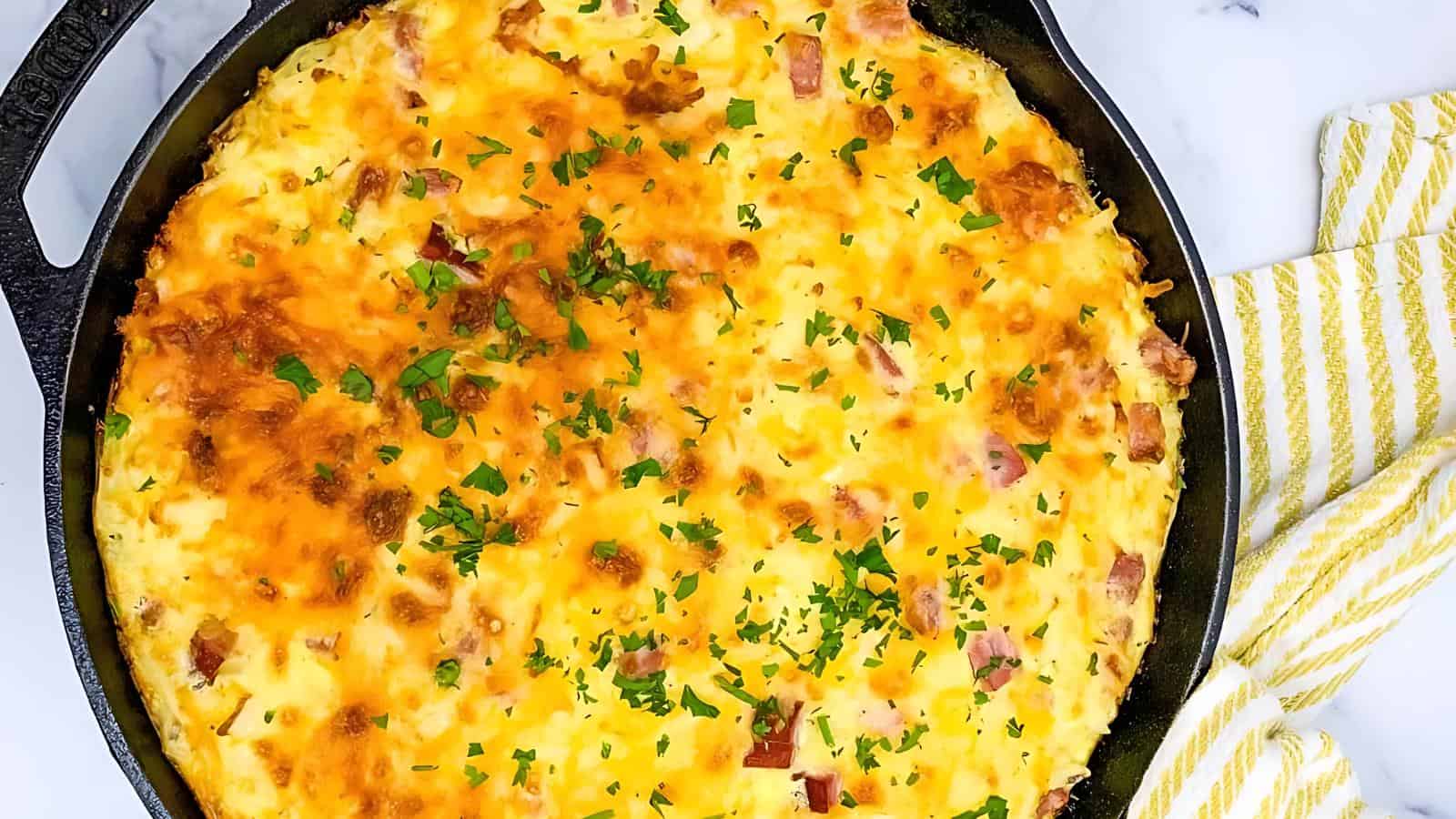A baked dish in a skillet topped with melted cheese and chopped herbs, garnished with parsley, placed on a striped yellow towel.