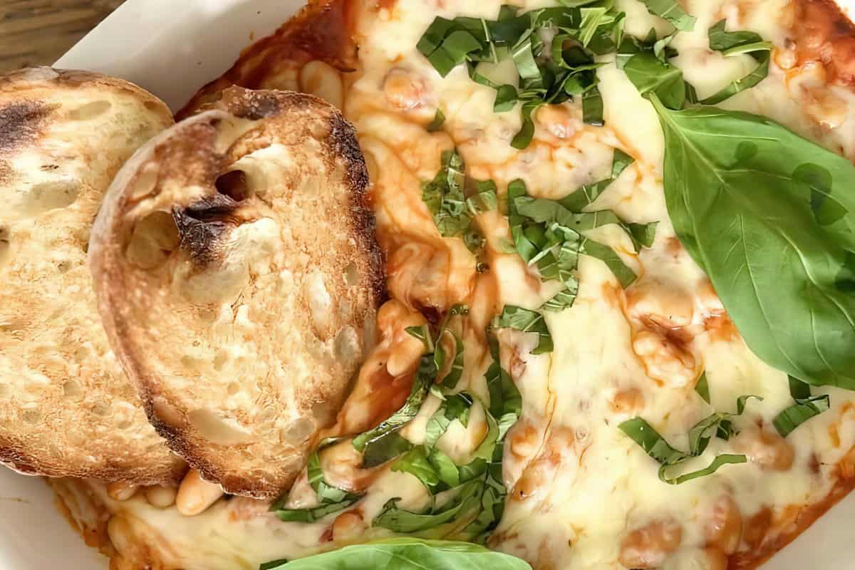 A close-up of a dish featuring melted cheese with chopped basil on top, accompanied by toasted slices of bread. The food is served in a white dish, with fresh green basil leaves as garnish.